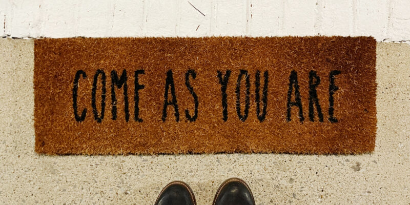 Person wearing black boots standing in front of a "Come As You Are" welcome mat.